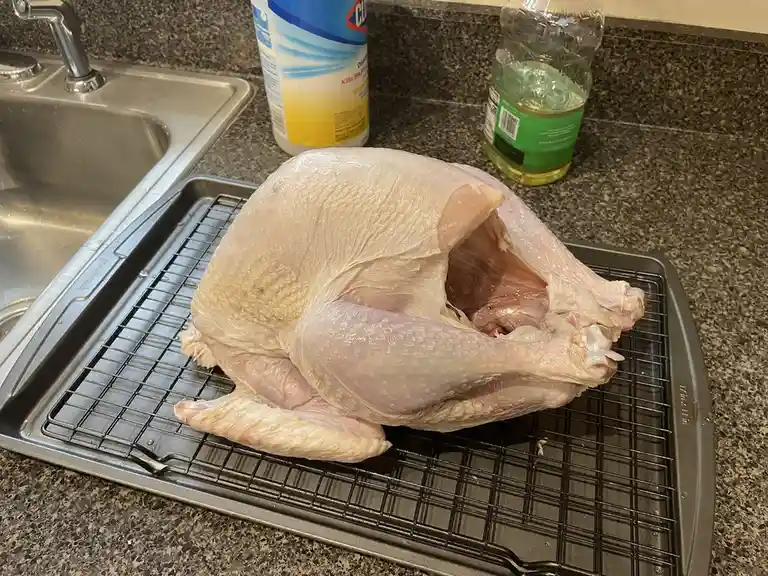 A raw, whole turkey sits on a metal roasting rack in a kitchen, its cavity open and facing upward. In the background, a…