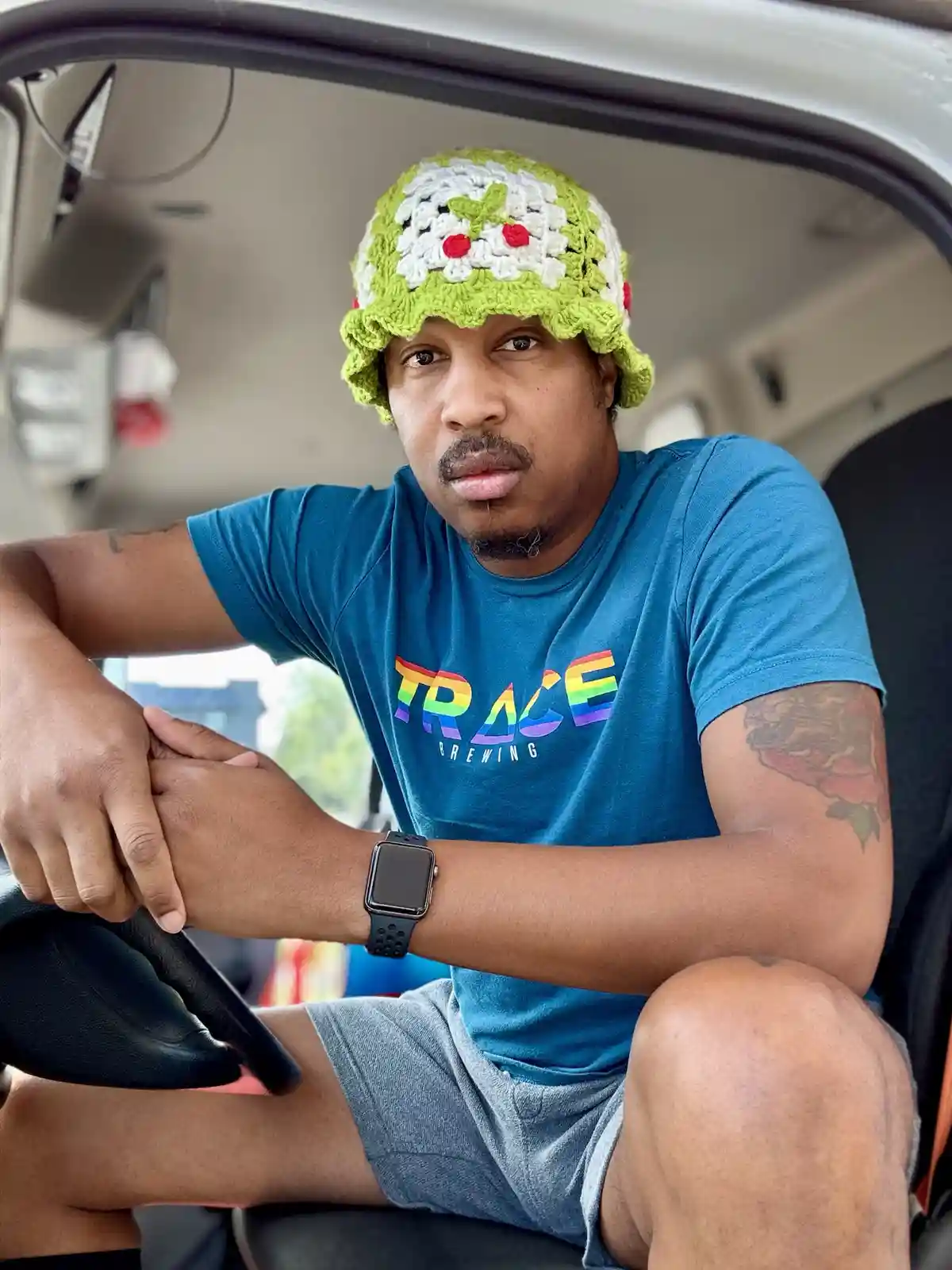 A man sits in the driver's seat of a vehicle, wearing a bright green and white crocheted hat with a flower design, looking…