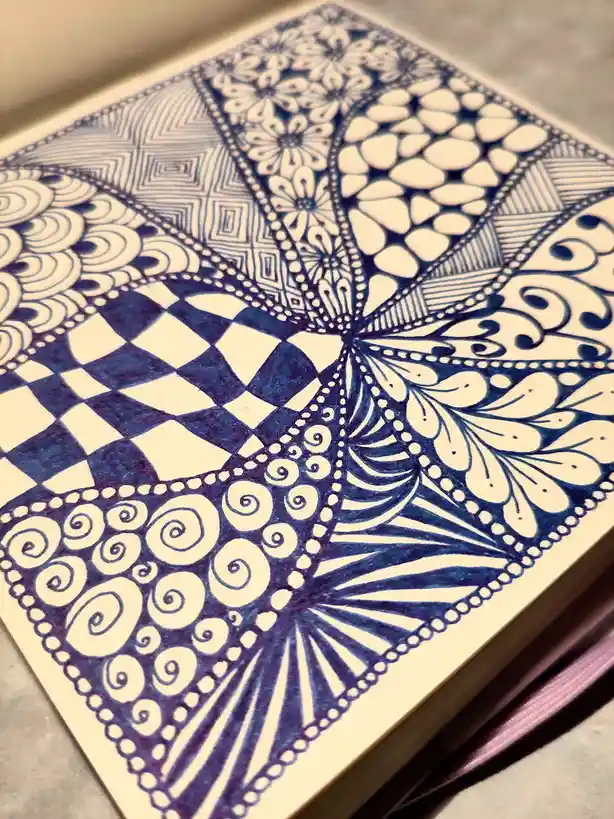 A close-up of a detailed, intricate zentangle-style drawing on paper, featuring symmetrical patterns including…