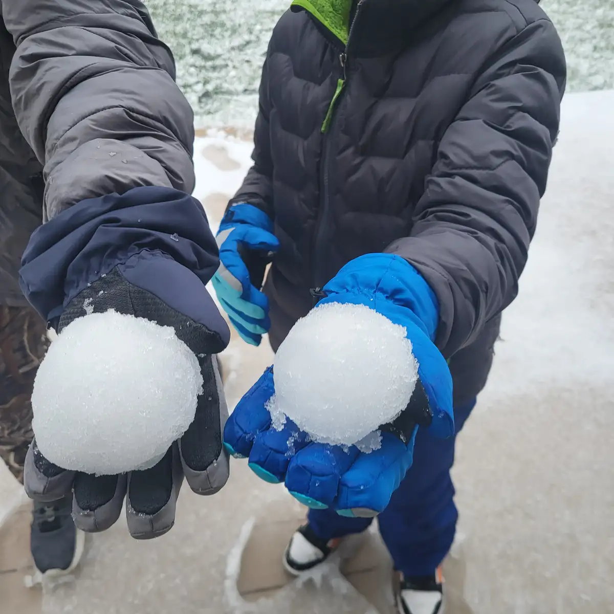 Two children in winter jackets and gloves hold up snowballs, one in dark gloves and one in blue gloves, comparing their…