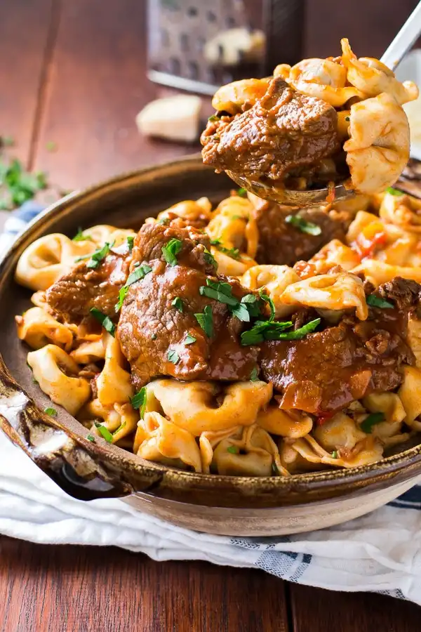A close-up photo of a hearty pasta dish in a ceramic bowl, featuring large, shell-shaped pasta mixed with chunks of meat in…