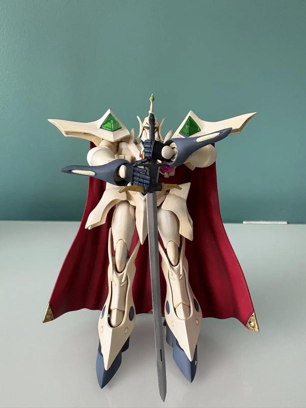 A custom-painted, unpainted white and grey mecha model with green triangular headpieces and a red cape stands holding a…
