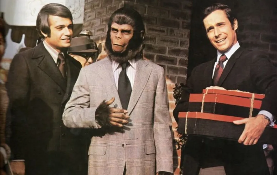three men in suits stand together, the central figure wearing a full ape costume with a human-like face, while the man on…