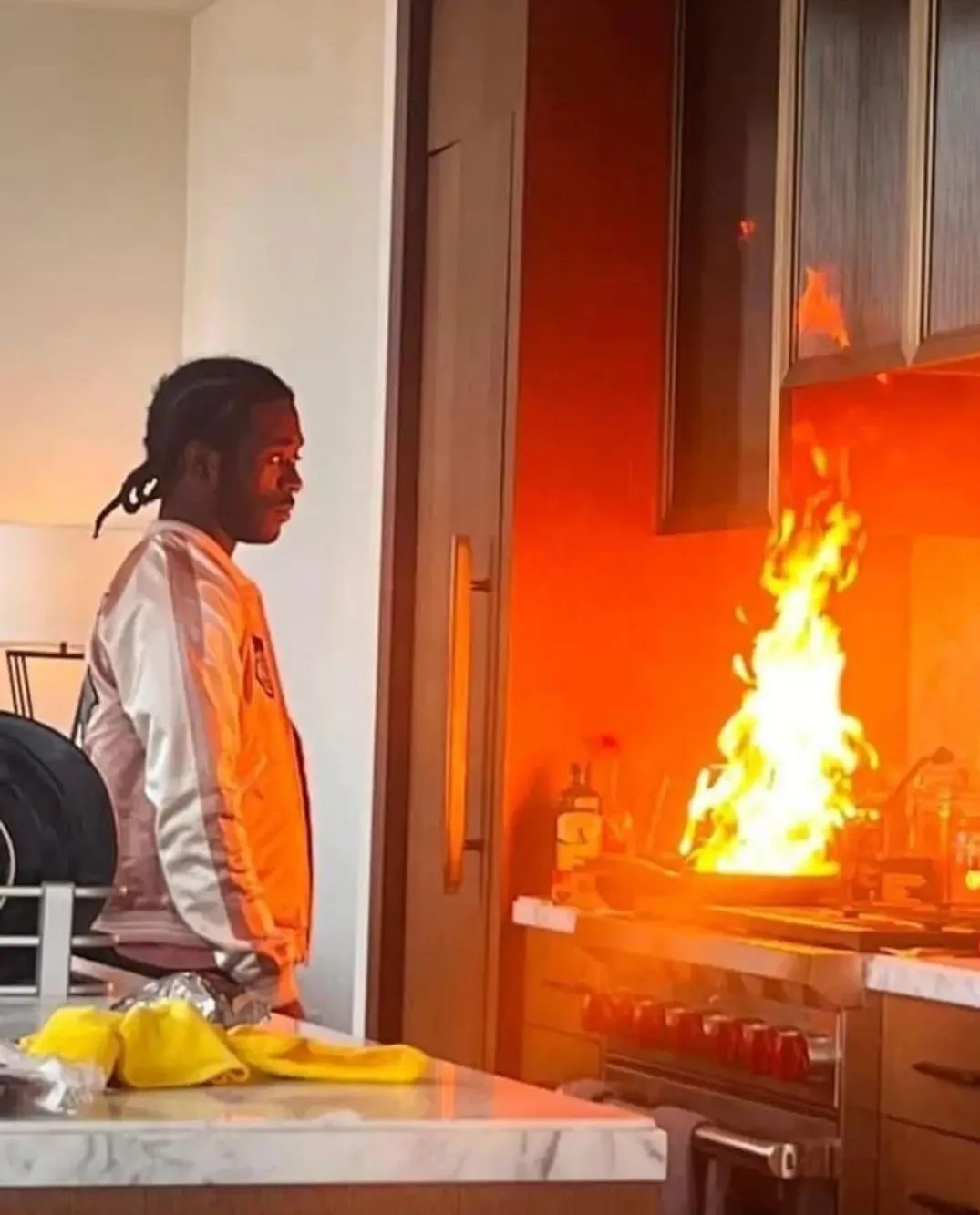 A man in a white and orange jacket stands in a modern kitchen, looking calmly at a large fire erupting from a stove burner…