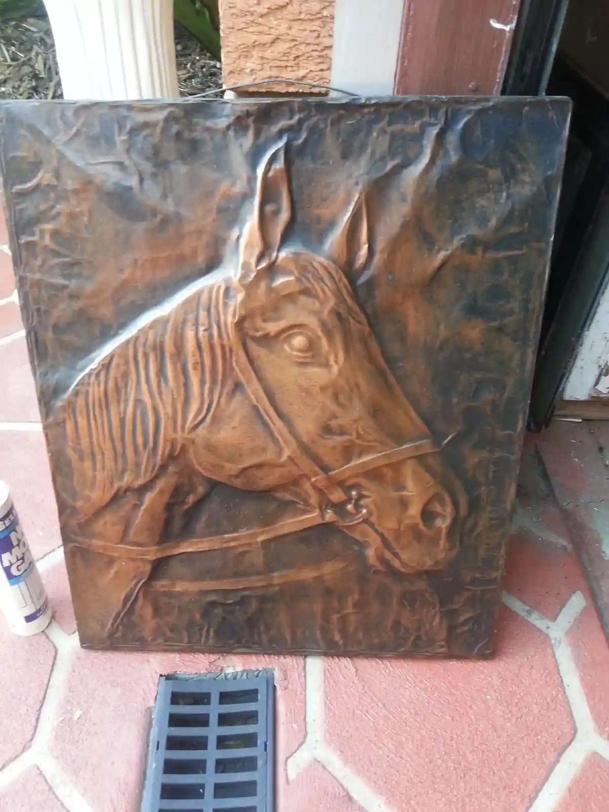 A bronze-colored metal relief sculpture of a horse's head and neck, leaning against a wall on a tiled patio. The horse wears…