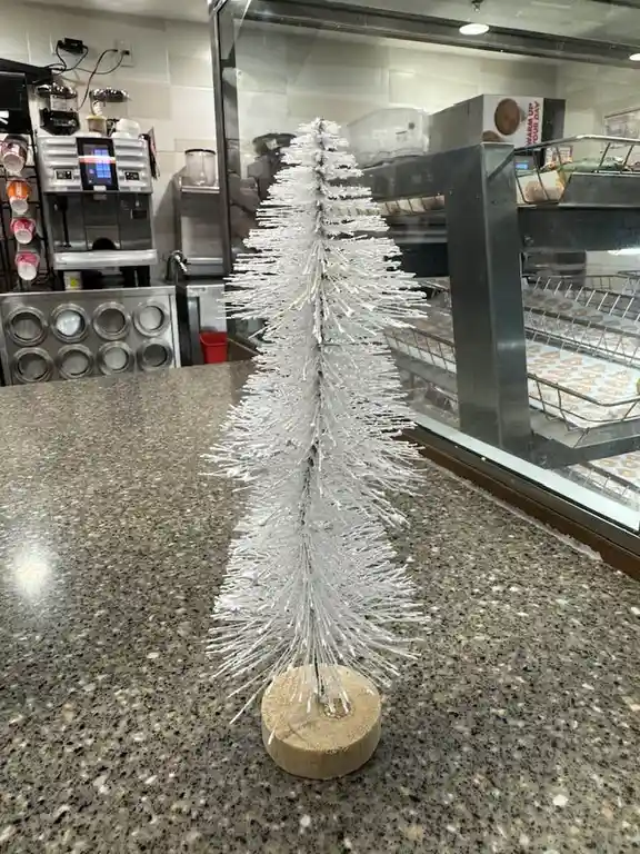 A small, white, bristle-covered tree on a wooden base sits on a speckled countertop in what appears to be a bakery or coffee…