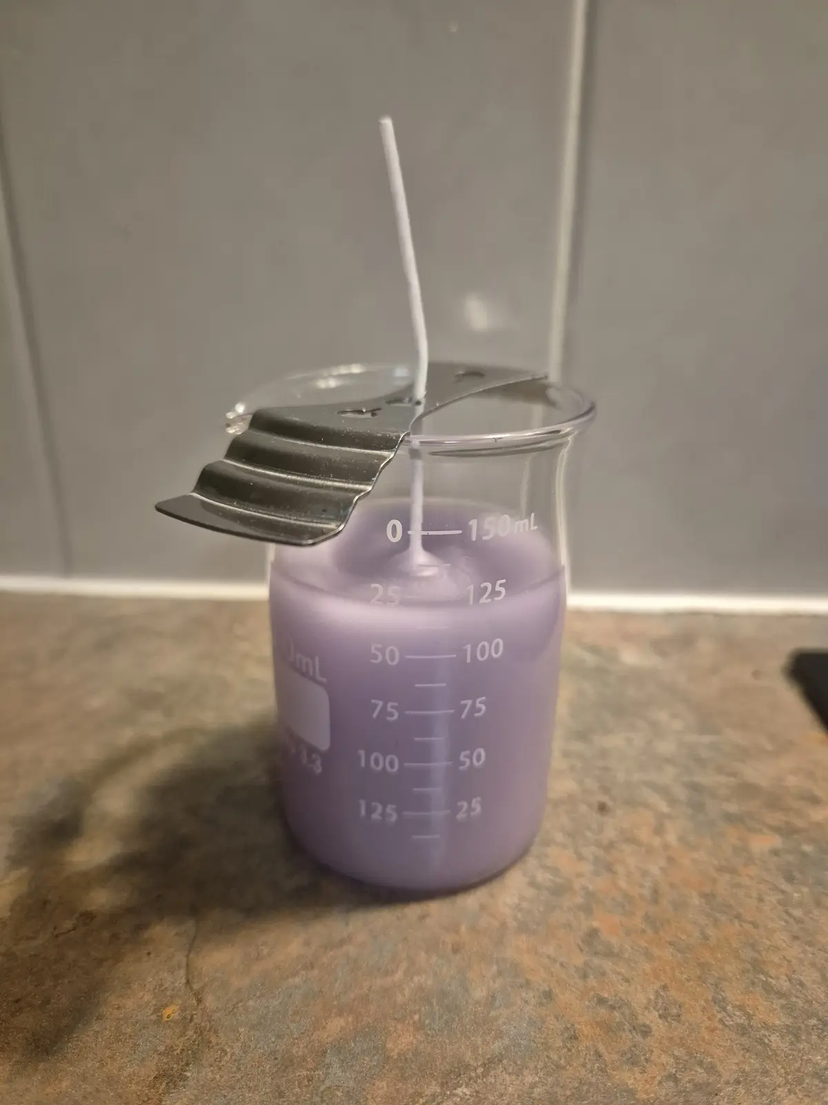 A purple candle wax is poured into a clear glass beaker marked with volume measurements up to 150mL, with a white wick…