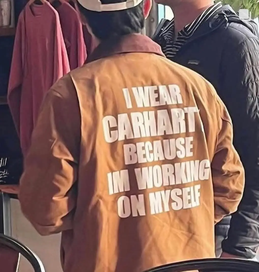 A person viewed from behind wears a brown Carhartt jacket with large white text on the back that reads 'I WEAR CARHARTT…