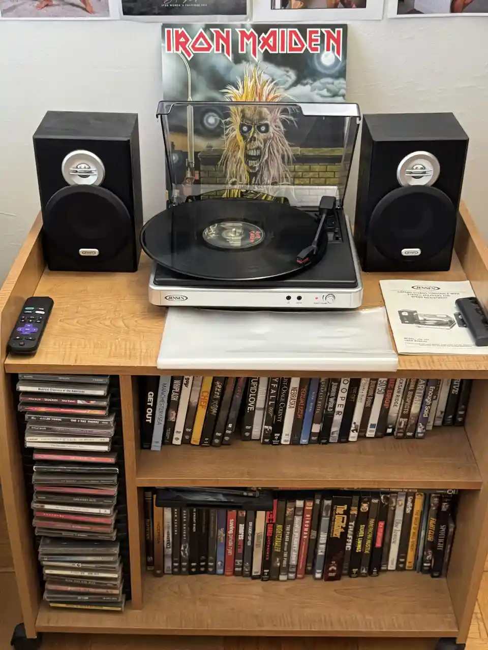 A wooden media cart holds a Jensen turntable with a vinyl record spinning, flanked by two black Jensen speakers. Behind the…
