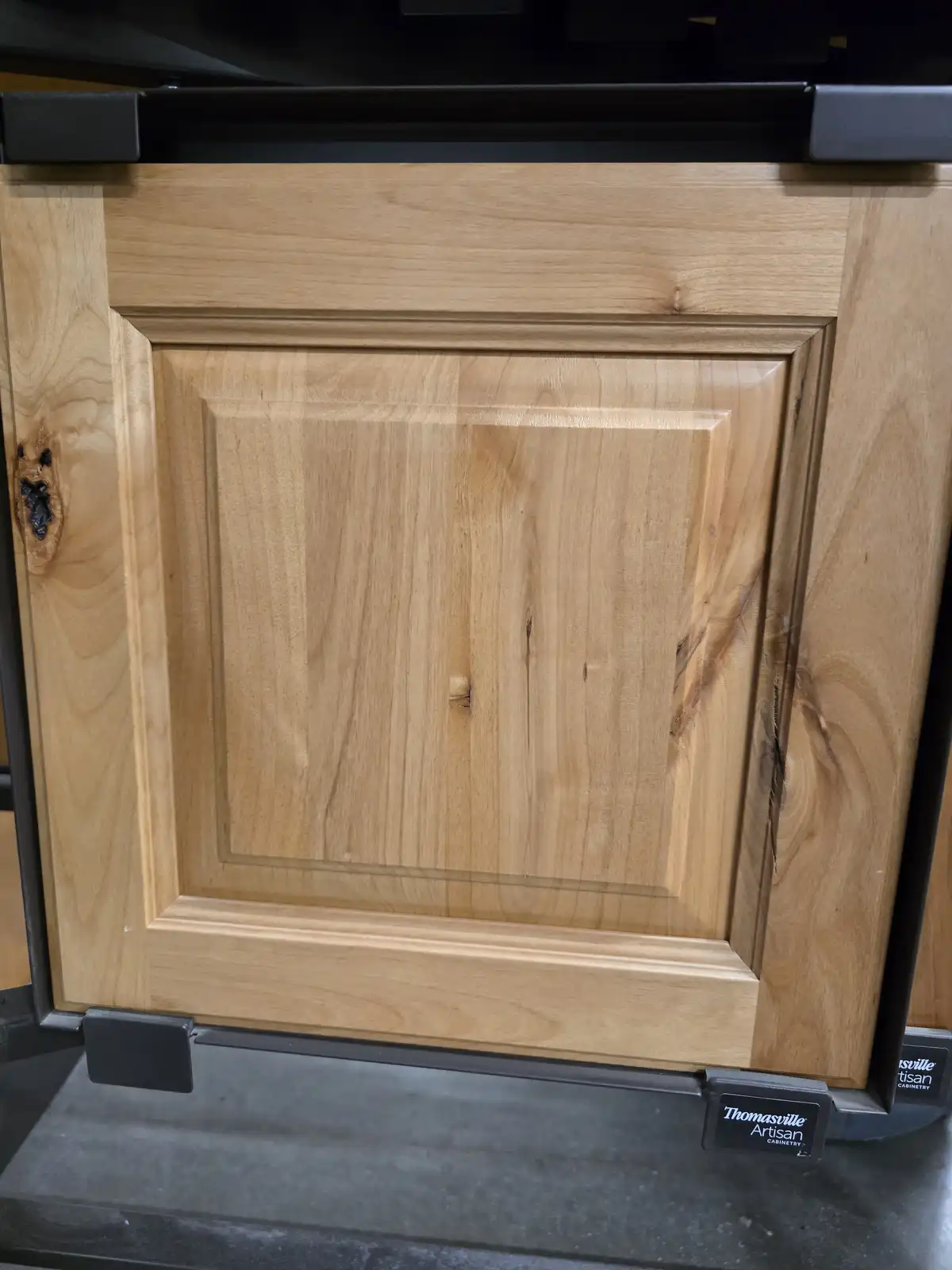 A close-up photo of a light-colored wooden cabinet door with a raised panel design, displayed in a showroom setting with…