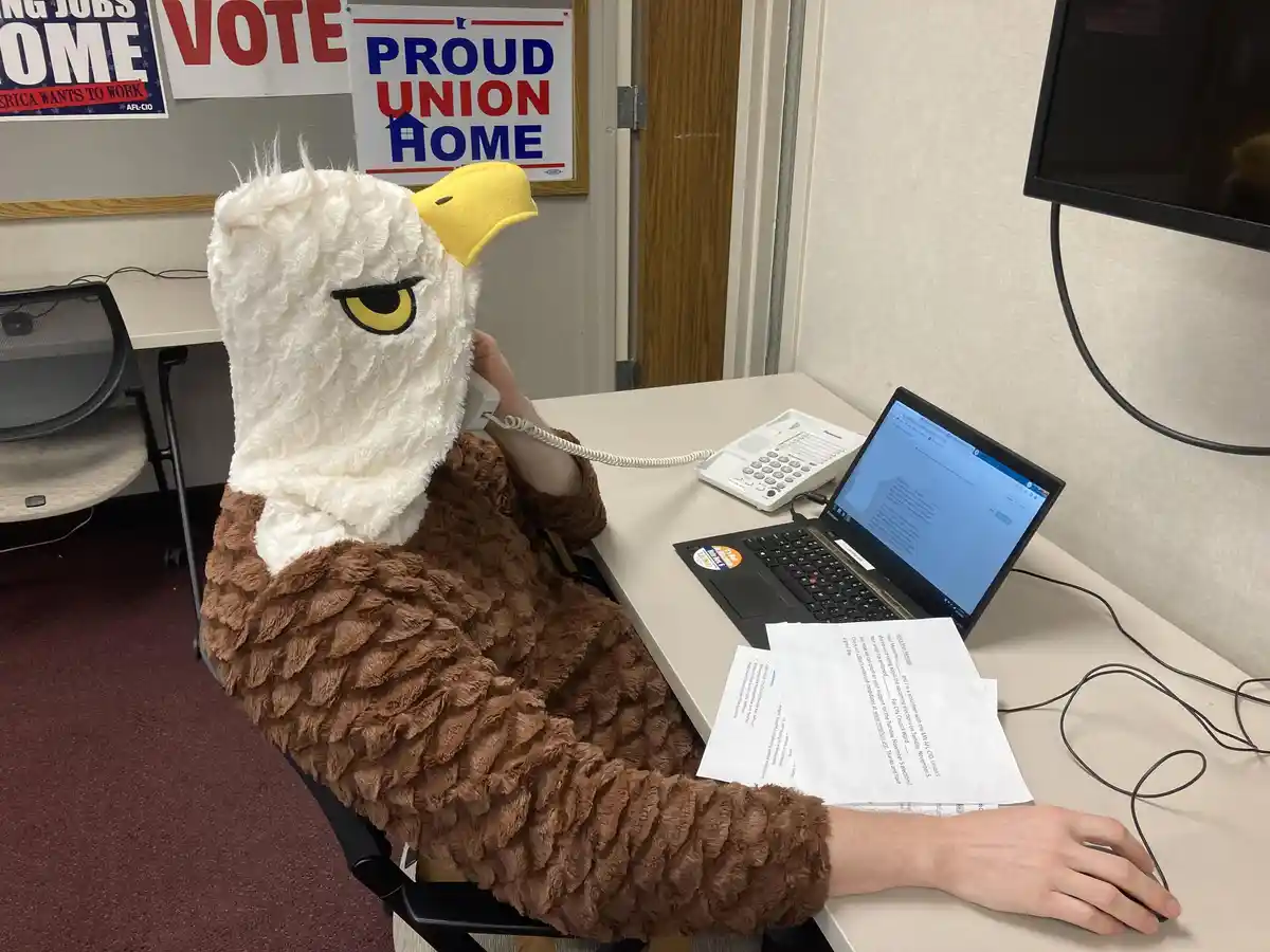 A person wearing a full-body, fluffy eagle costume sits at a desk in an office, holding a landline phone to their ear while…