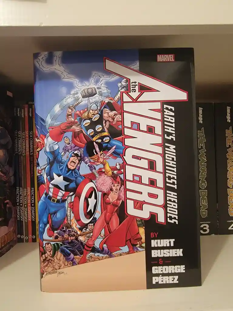 A comic book titled 'The Avengers: Earth's Mightiest Heroes' by Kurt Busiek & George Pérez sits on a bookshelf, featuring a…