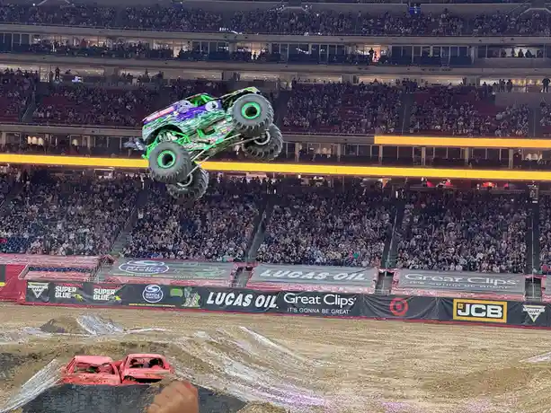 A green and purple monster truck is airborne mid-jump in a packed indoor arena, with a crowd of spectators in the stands…
