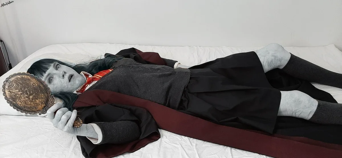 A person in a school uniform lies on a bed holding an ornate hand mirror, their face and hands painted pale gray with dark…