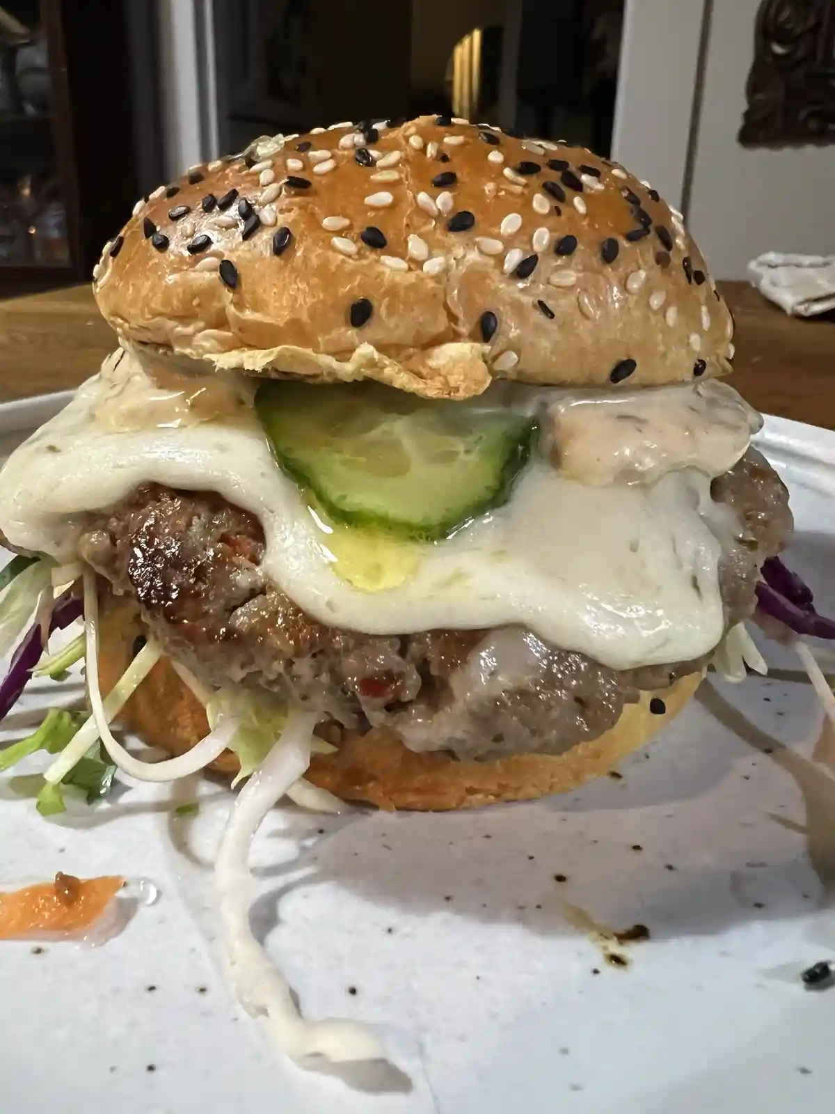 A close-up photo of a messy, overflowing burger on a white plate, featuring a sesame seed bun, a meat patty, a fried egg, a…