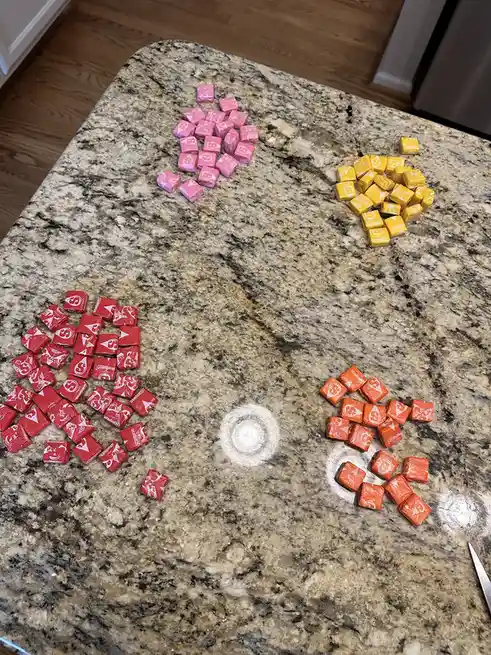 Four piles of Skittles candies arranged on a granite countertop to form the shape of a face: pink candies for the eyes…