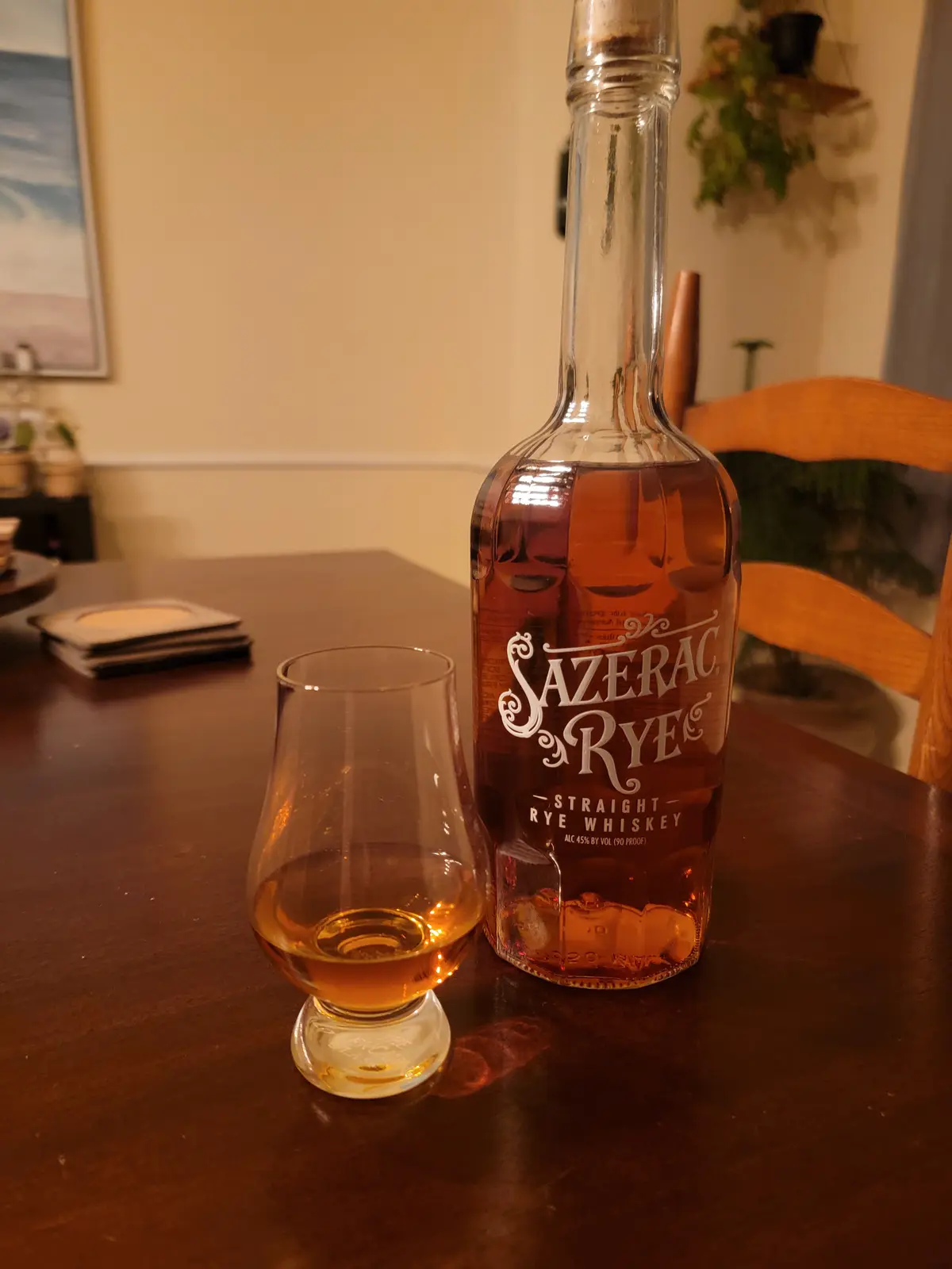 A bottle of Sazerac Rye Straight Rye Whiskey sits next to a tasting glass with a small amount of amber liquid on a dark…