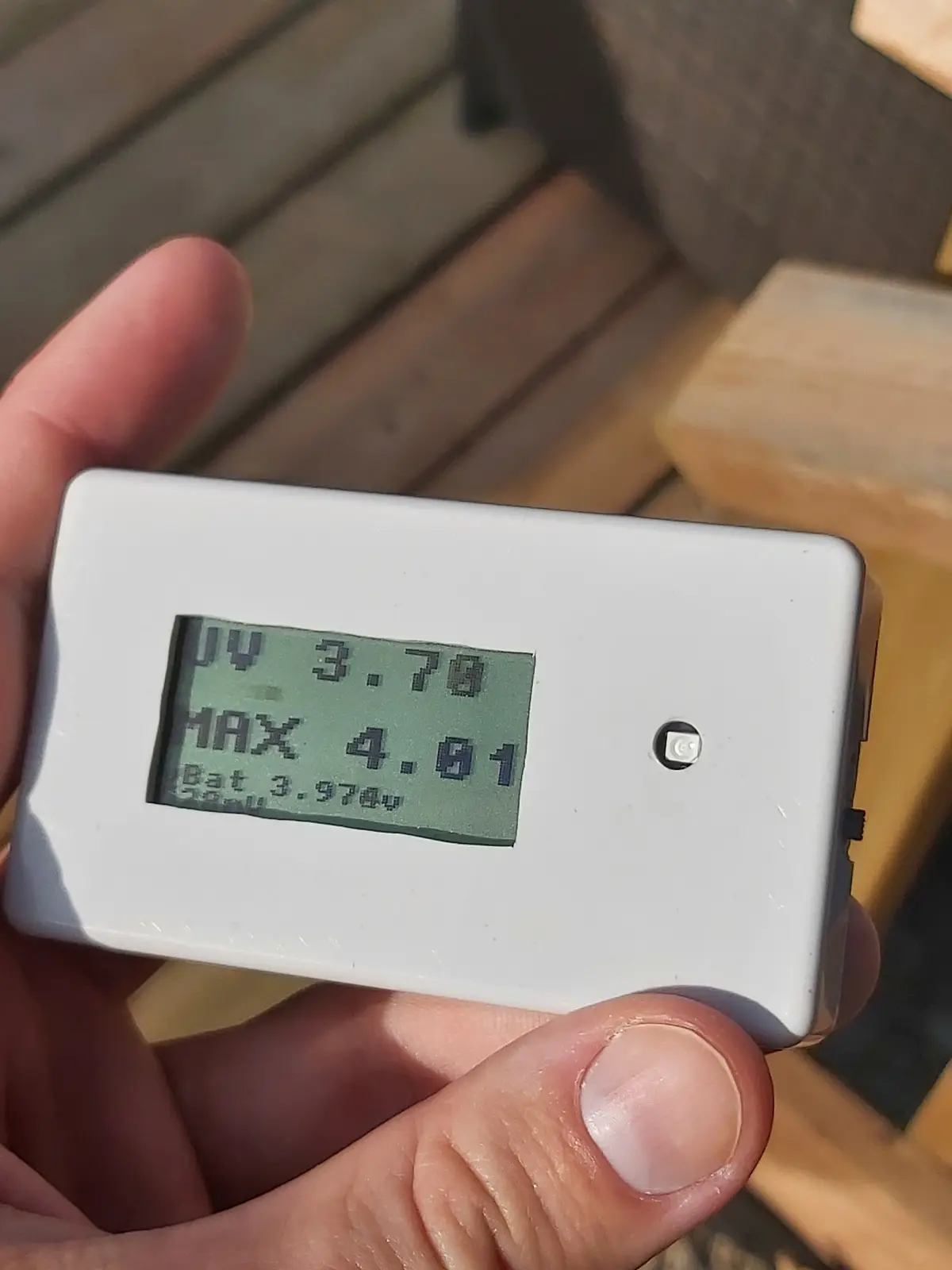 A hand holds a white handheld UV meter outdoors on a wooden deck, its small green LCD screen displaying 'UV 3.70', 'MAX…