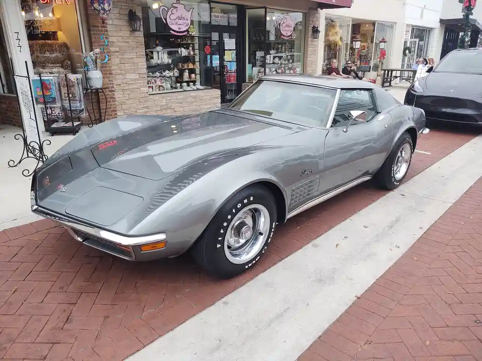 A silver classic Chevrolet Corvette Stingray is parked on a brick sidewalk in front of a tea shop. The car has chrome wheels…