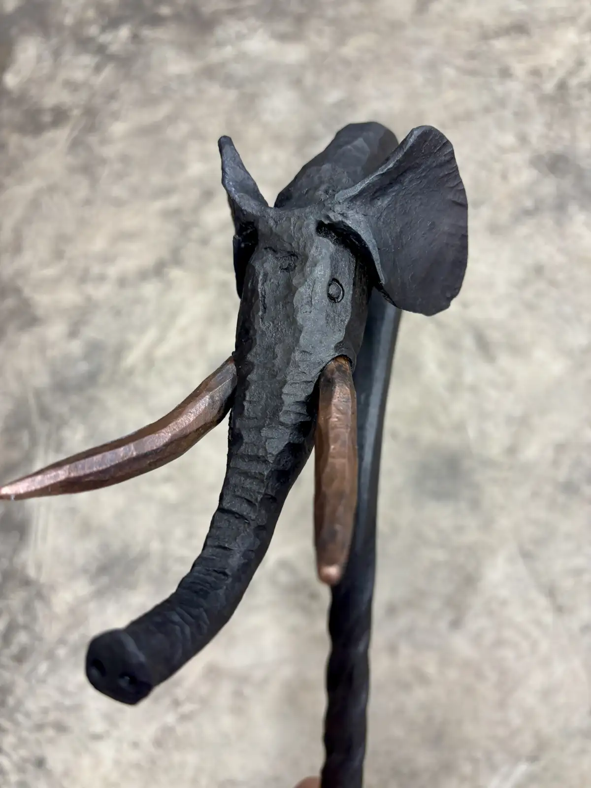 A close-up of a handcrafted metal sculpture of an elephant's head, featuring a dark, textured body and copper-colored tusks…