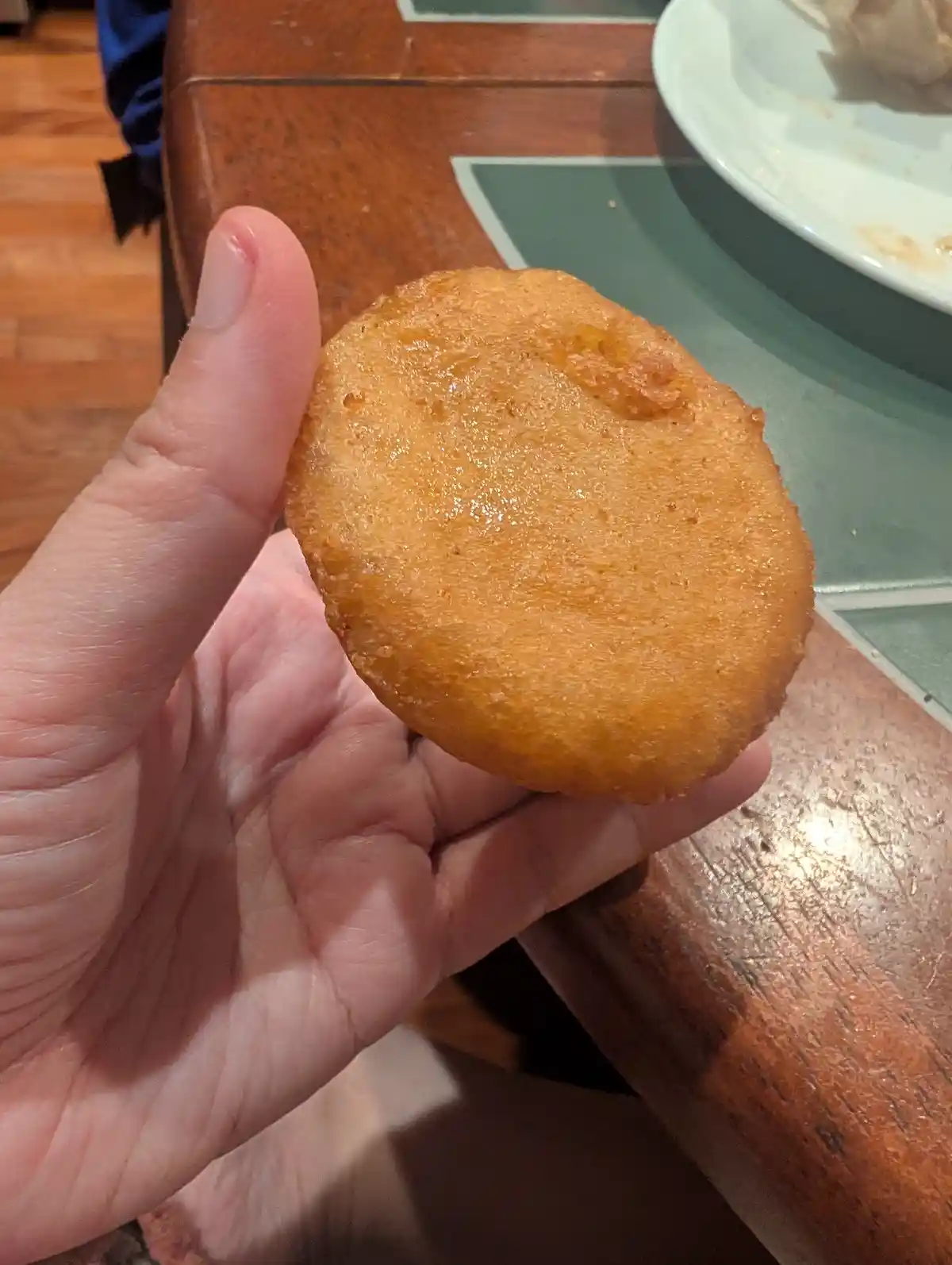 A hand holds up a golden-brown, round, fried hush puppy against a wooden table with a green placemat and a partially visible…