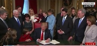 A man in a red tie sits at a desk signing a document while surrounded by people, including children. A small child in a red…