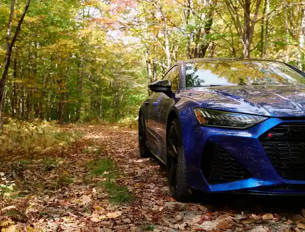A shiny blue Audi RS7 sports car is parked on a leaf-covered dirt path in a dense autumn forest, with trees displaying…