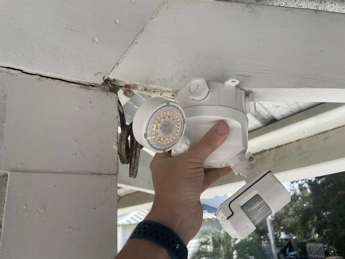A hand holds a white outdoor motion-sensor light fixture mounted under a porch roof, with visible wiring and a blue…