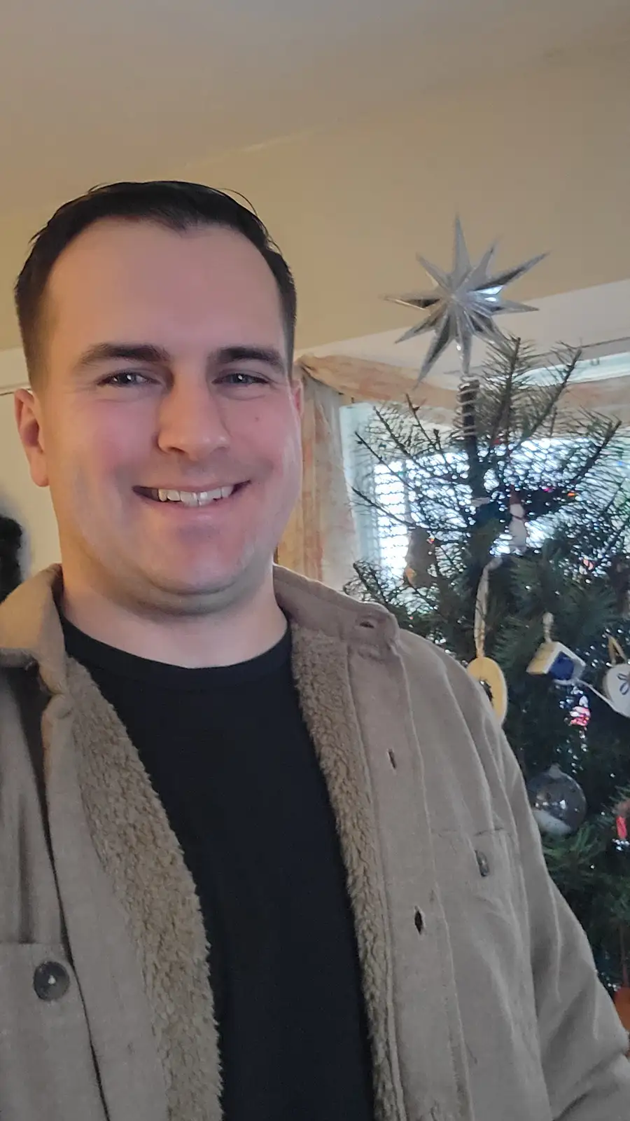 A smiling man in a beige sherpa-lined jacket over a black shirt poses for a selfie in front of a decorated christmas tree…