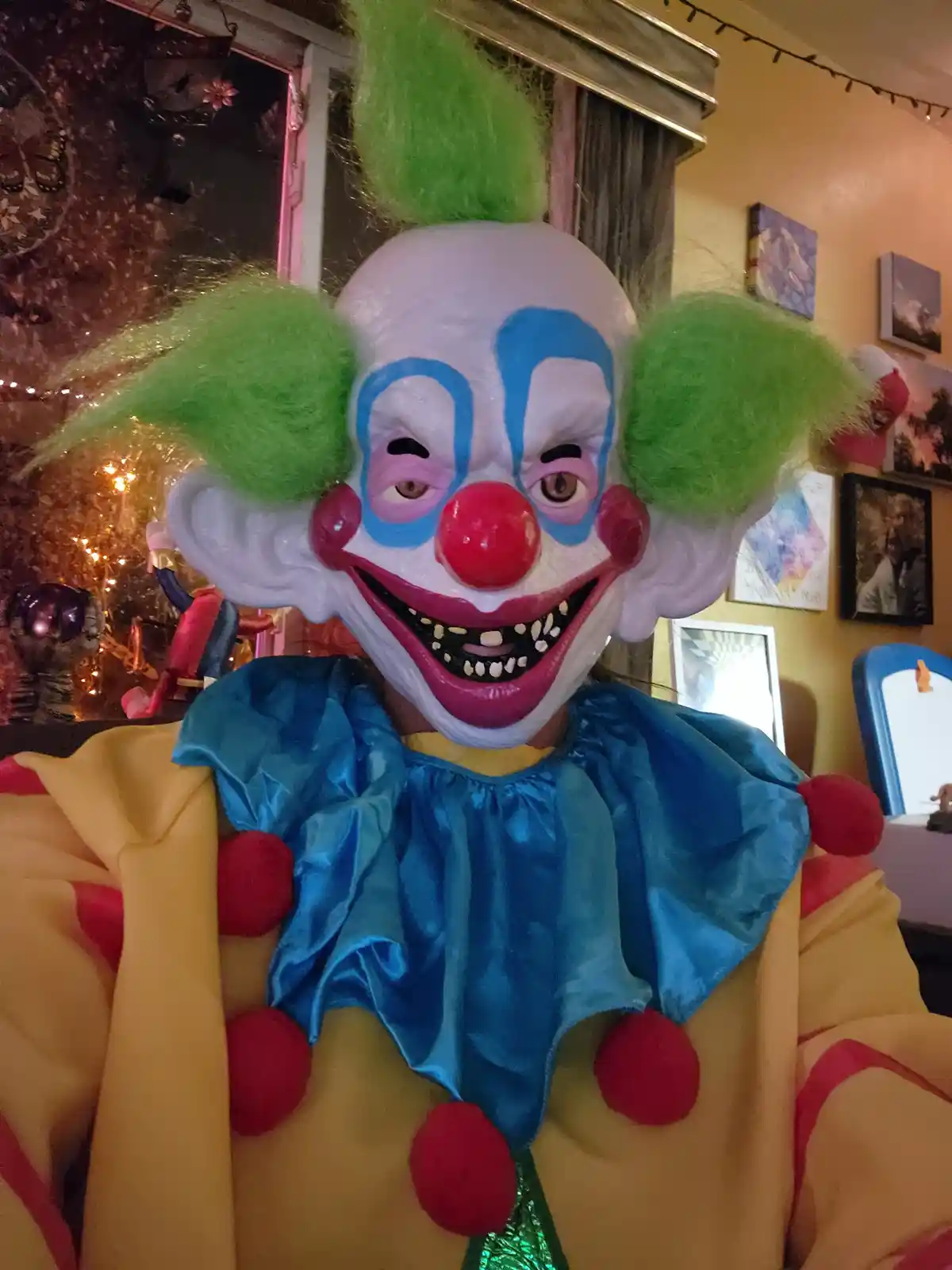 A person wearing a scary clown mask with green hair, blue makeup, and a wide grin with visible teeth, dressed in a yellow…