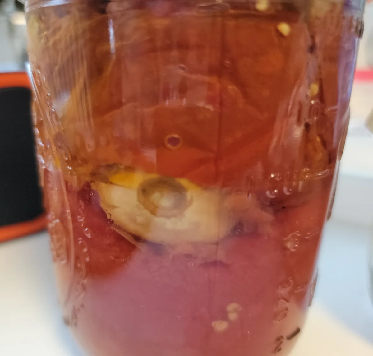 A close-up photo shows a glass mason jar filled with a reddish-brown liquid, containing a pickle and other pickling…