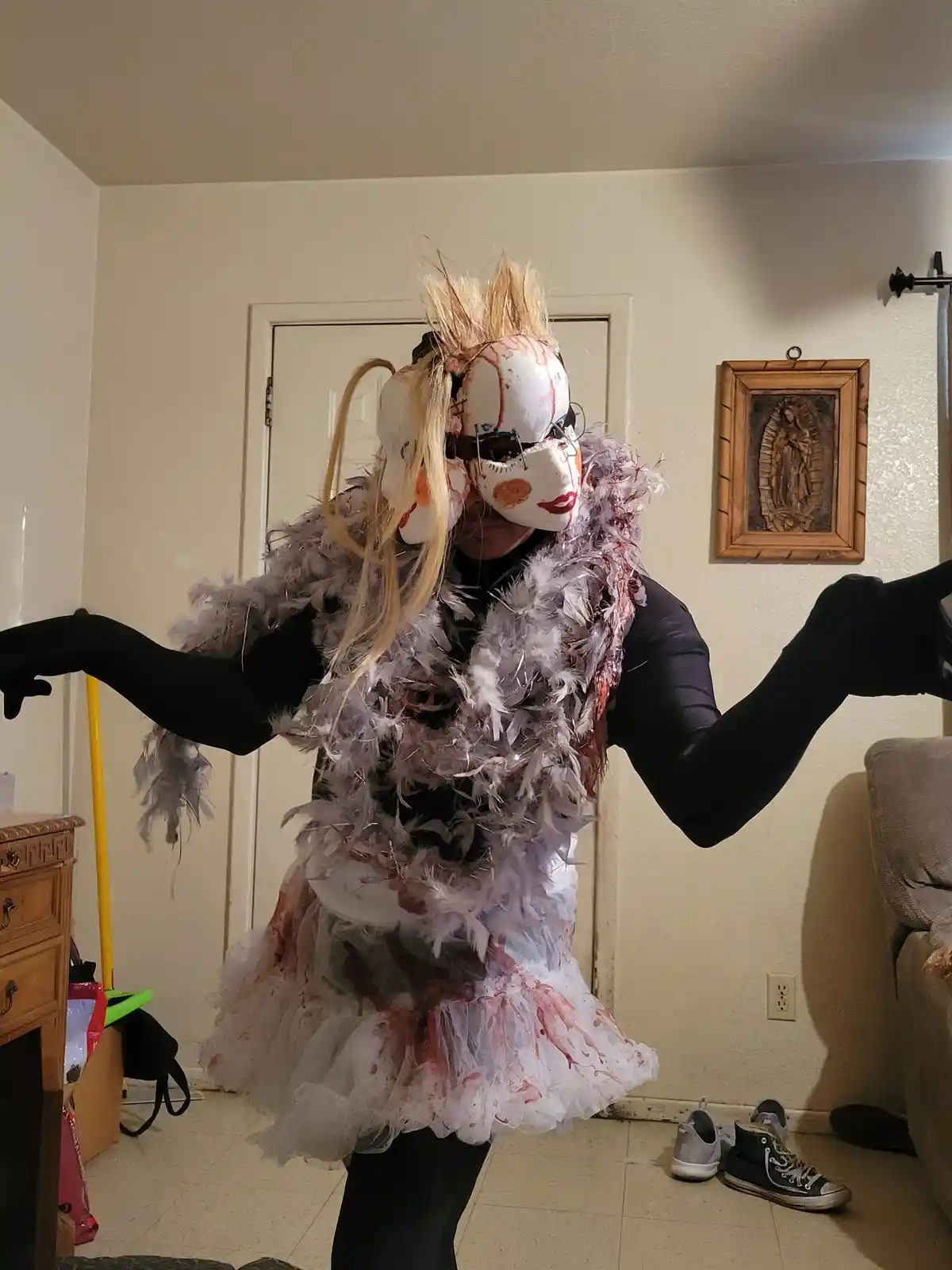 A person in a homemade costume stands in a room with arms outstretched, wearing multiple white Venetian-style masks with red…