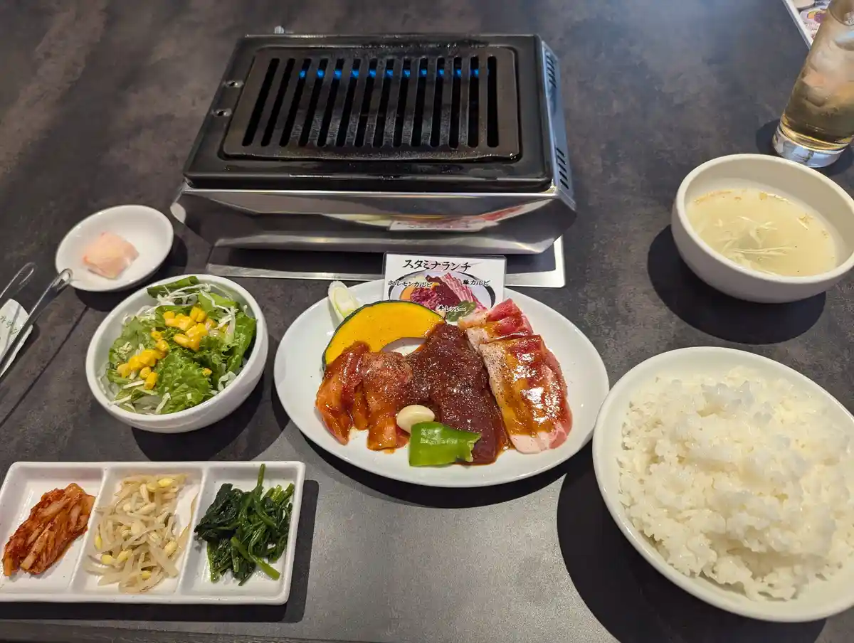 A table set with a Korean BBQ meal featuring a built-in grill with blue flames, a plate of marinated meats and vegetables, a…