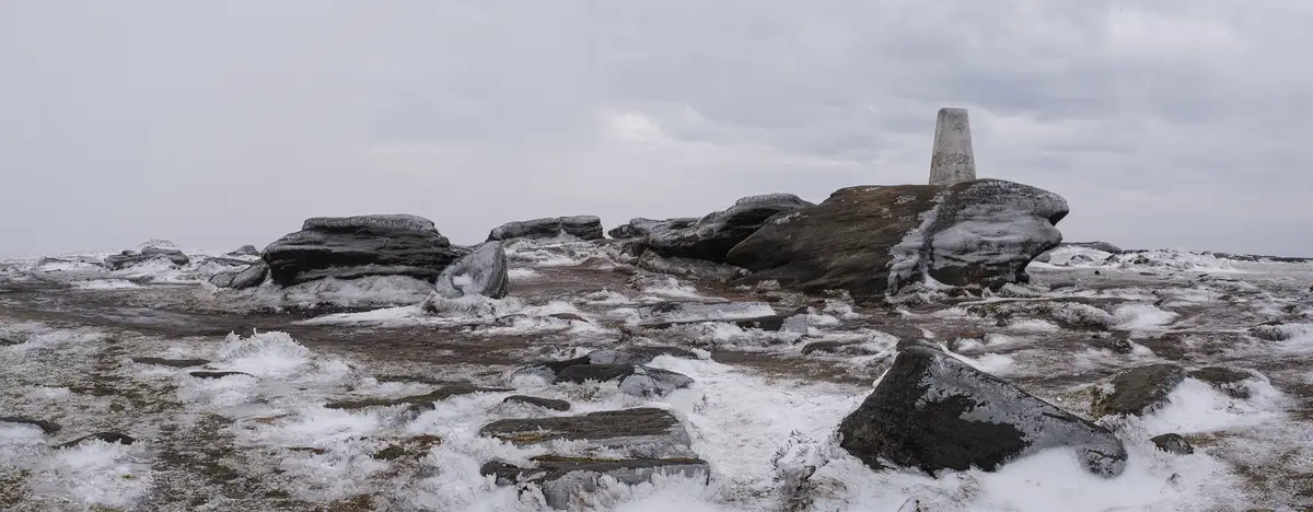 A wide, desolate landscape of frost-covered rocks under a gray sky, with a concrete trig point on a large boulder in the…