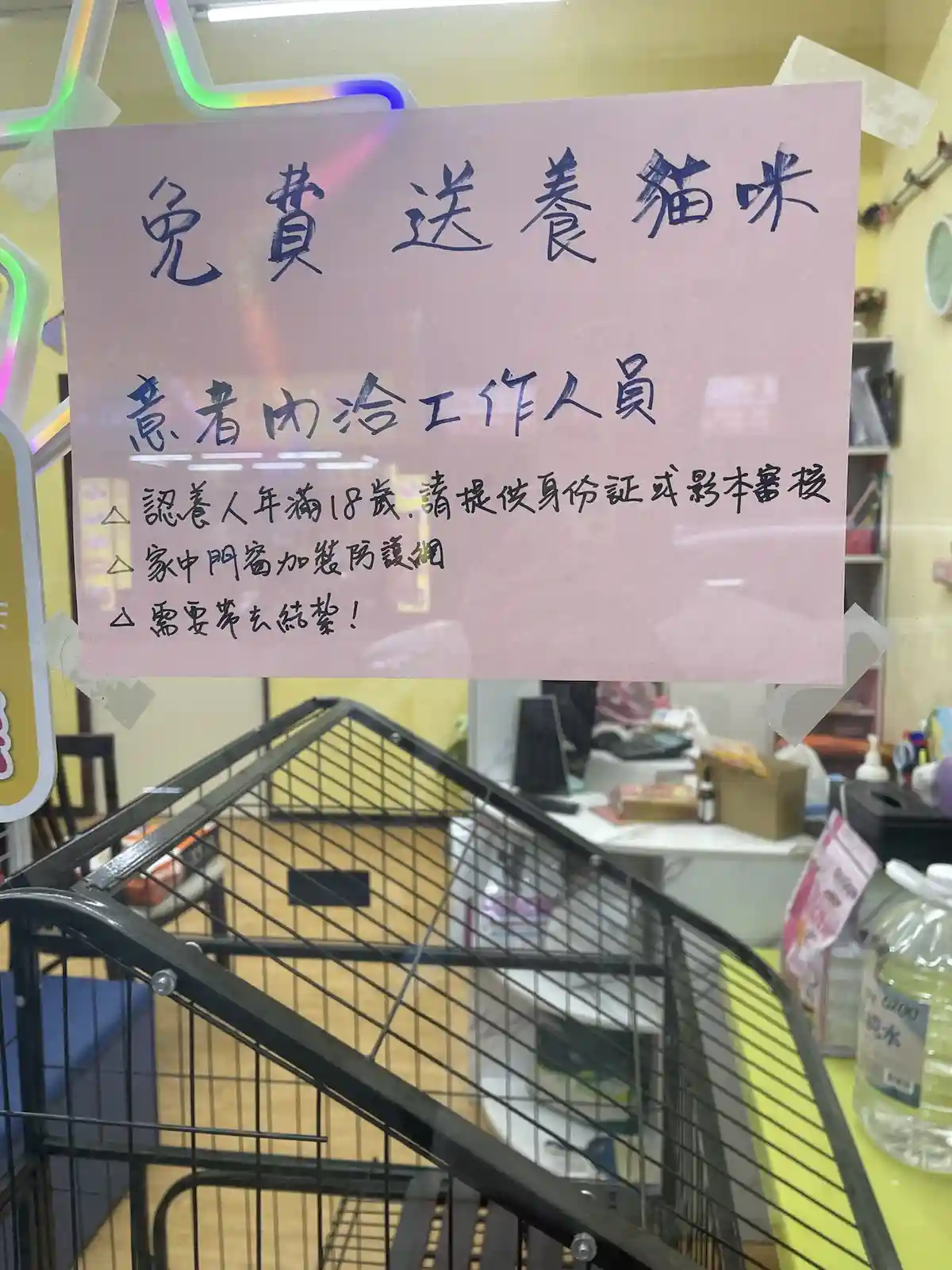 A handwritten pink sign taped to a glass door in a cat shelter or adoption center, advertising free cat adoption with strict…