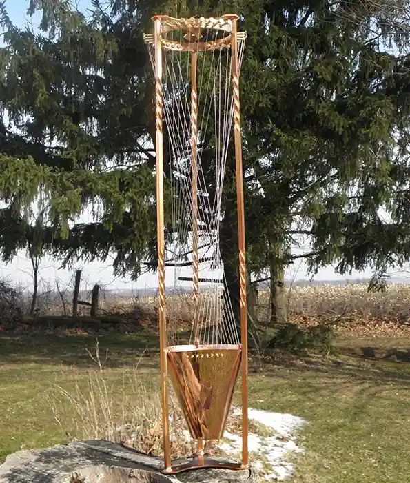 A tall, ornate, copper-colored harp sculpture stands outdoors on a stone base, surrounded by grass, trees, and patches of…