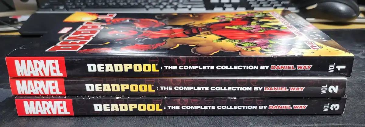 Three stacked Marvel comic books titled 'Deadpool: The Complete Collection by Daniel Way', volumes 1, 2, and 3, sit on a…