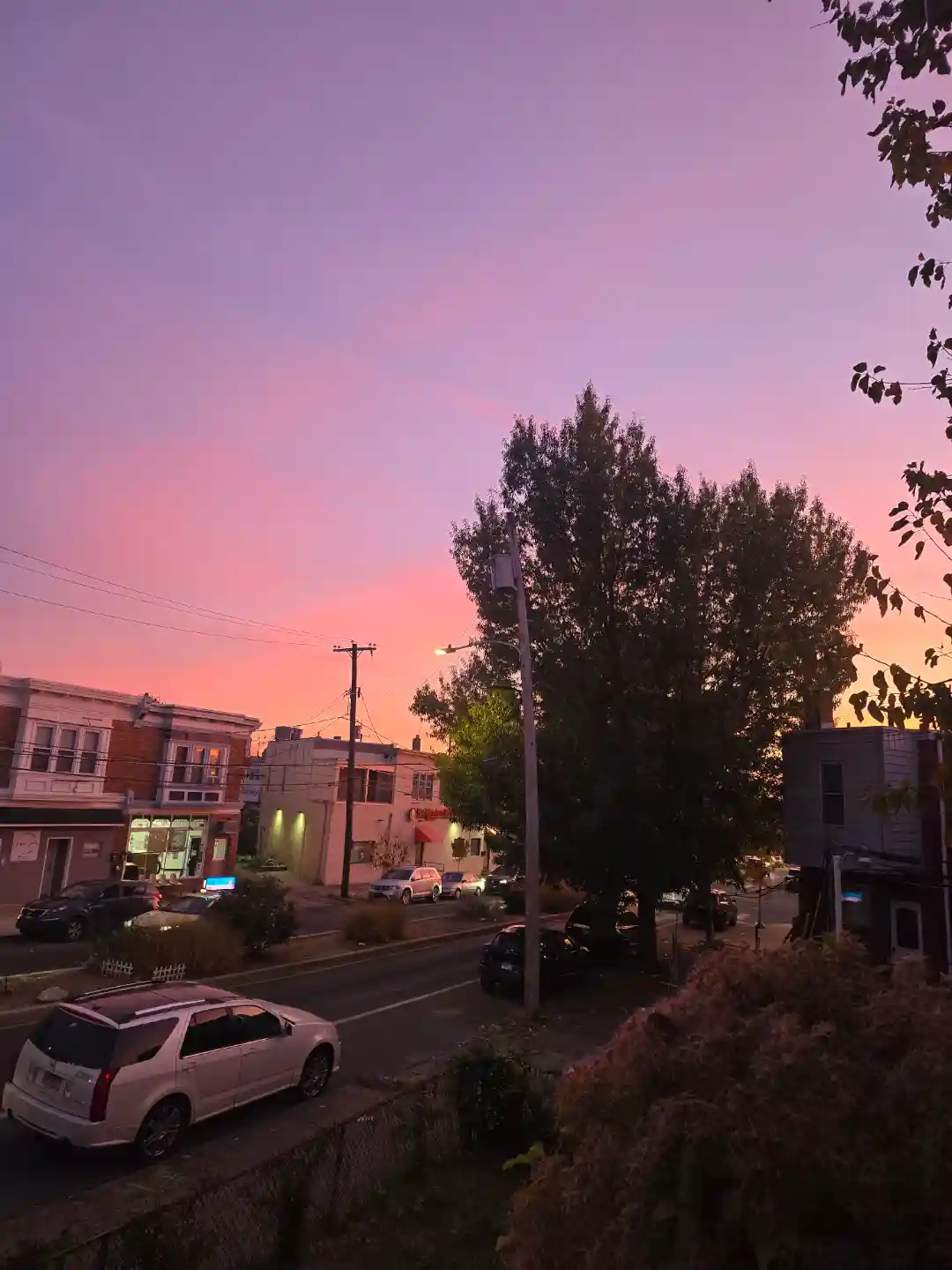 A photograph of a residential street at sunset, showing a vibrant pink and purple sky above parked cars, trees, and brick…