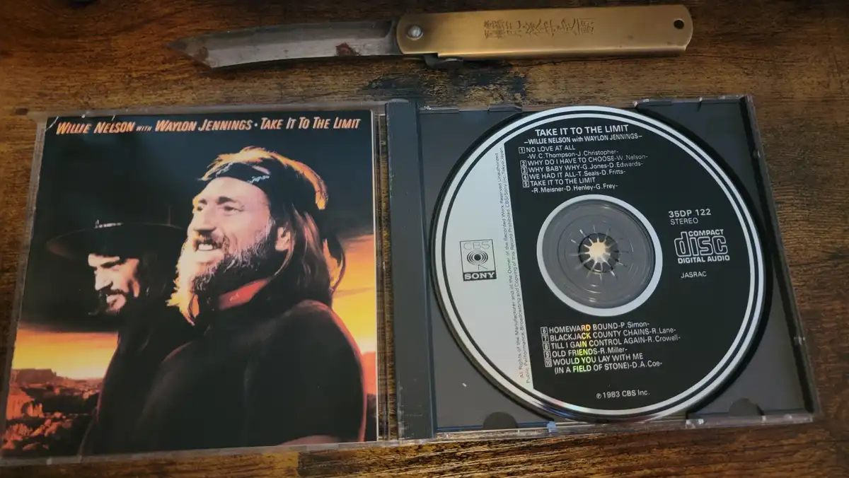 An open CD case for Willie Nelson with Waylon Jennings' 1983 album 'Take It To The Limit' sits on a wooden surface, with a…