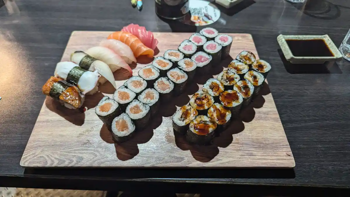 A wooden platter holds an elaborate sushi arrangement shaped like the letter 'K', featuring various rolls and nigiri, with…