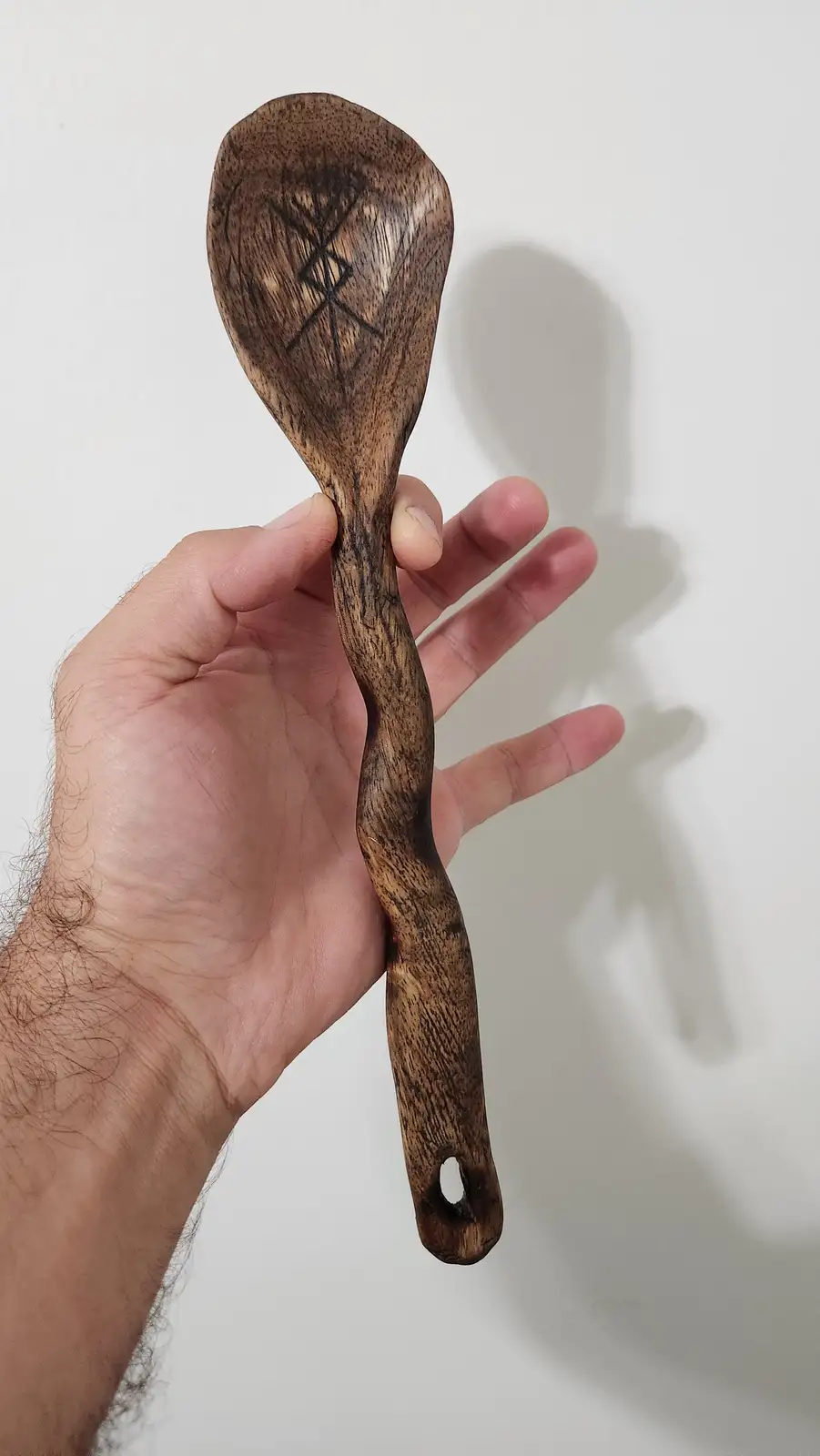 a hand holds a dark brown wooden spoon against a plain white background; the spoon's bowl features a carved symbol…