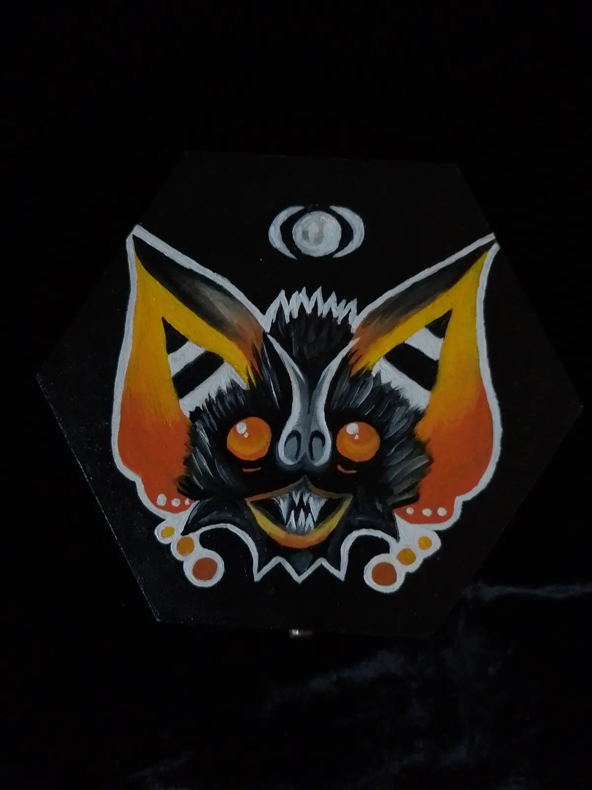 A hand-painted hexagonal box features a stylized demonic face with glowing orange eyes, sharp teeth, and large bat-like ears…