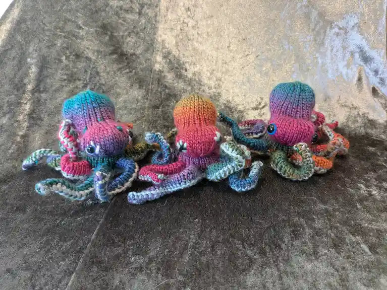 Three small, colorful knitted octopuses with googly eyes and multi-hued tentacles are arranged side-by-side on a textured…