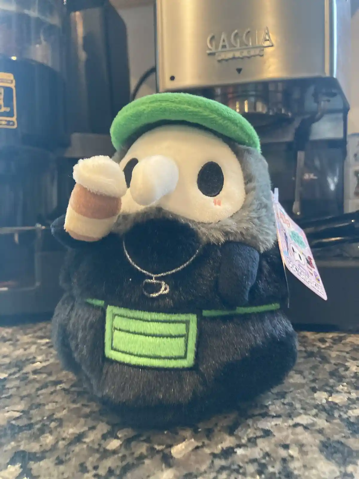 A plush toy resembling a skeletal coffee shop worker with a green hat and apron, holding a coffee cup, sits on a granite…