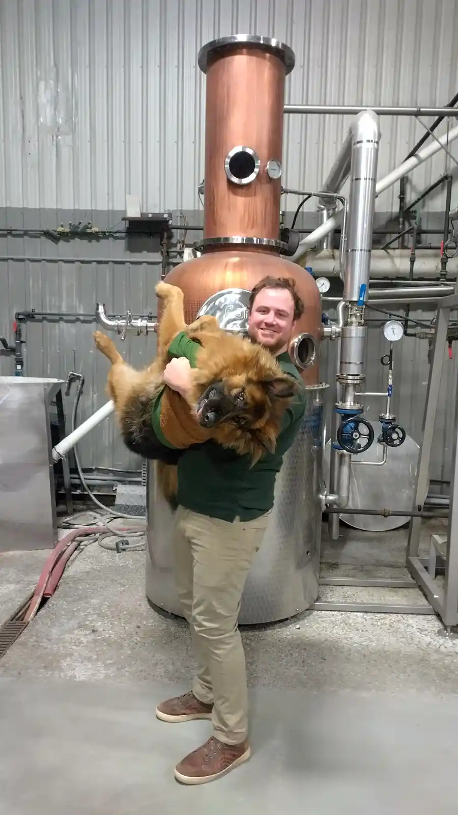 A man in a green shirt and khaki pants stands in front of a large copper and stainless steel distillation apparatus, smiling…