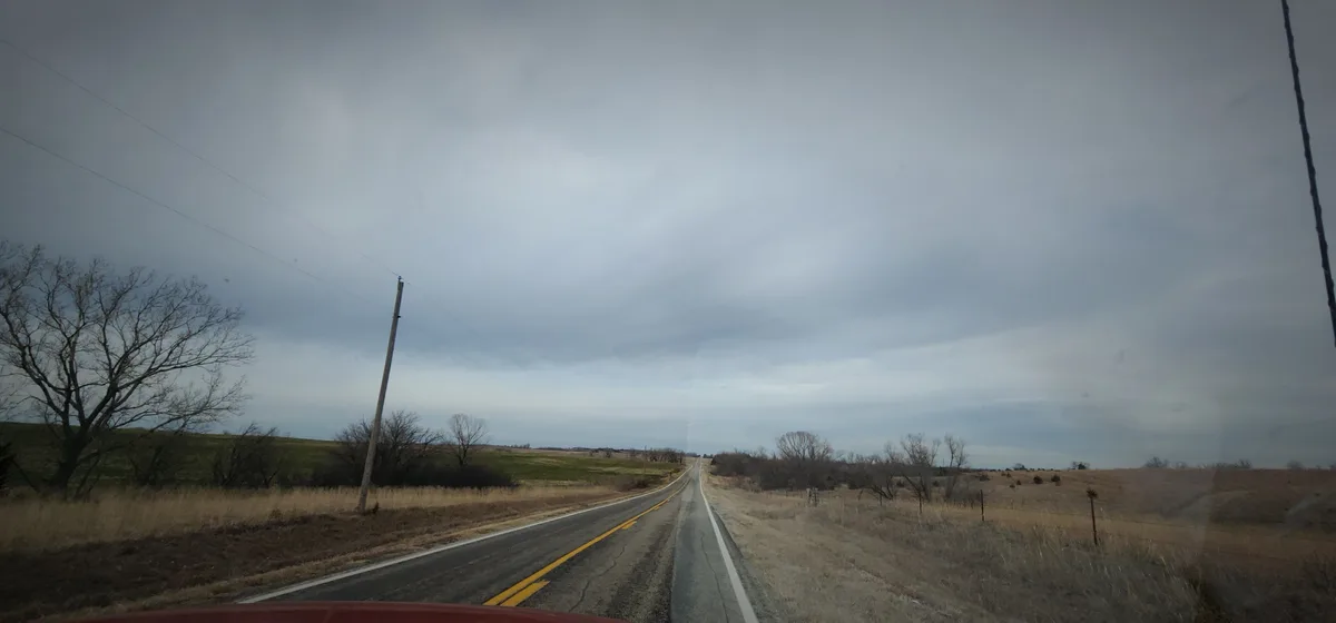 A photo taken from inside a car shows a long, straight, rural road stretching into the distance under an overcast sky. The…