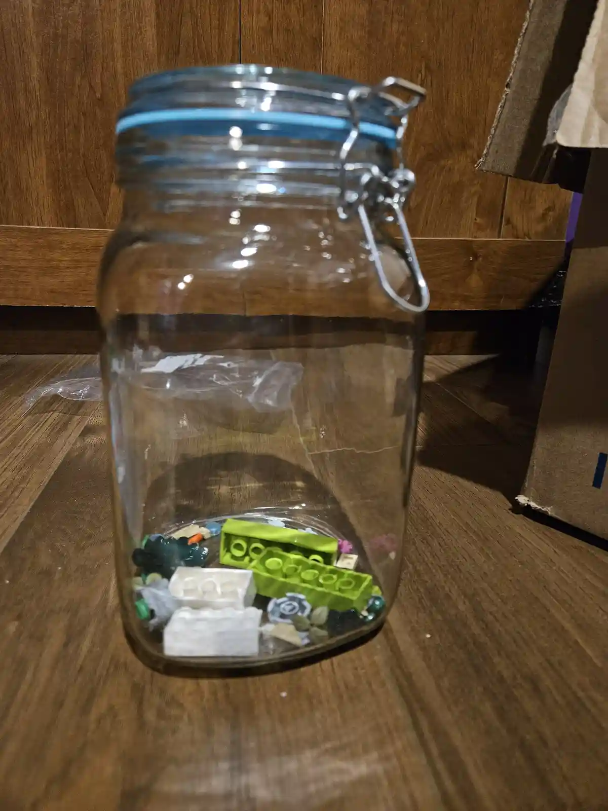 A clear glass jar with a metal clamp lid sits on a wooden floor, containing only a small amount of scattered Lego bricks at…