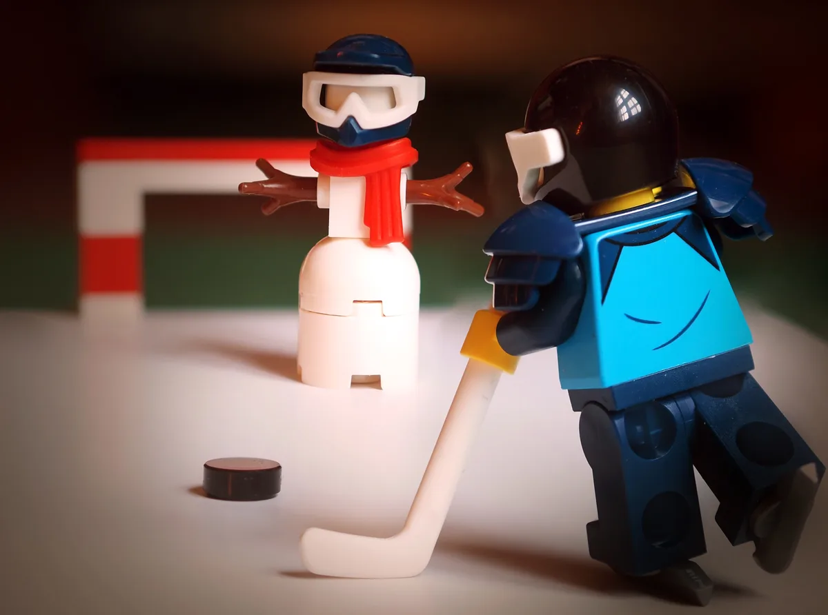 A Lego minifigure dressed as a hockey goalie holds a stick near a snowman made of Lego pieces, with a puck on the ground…