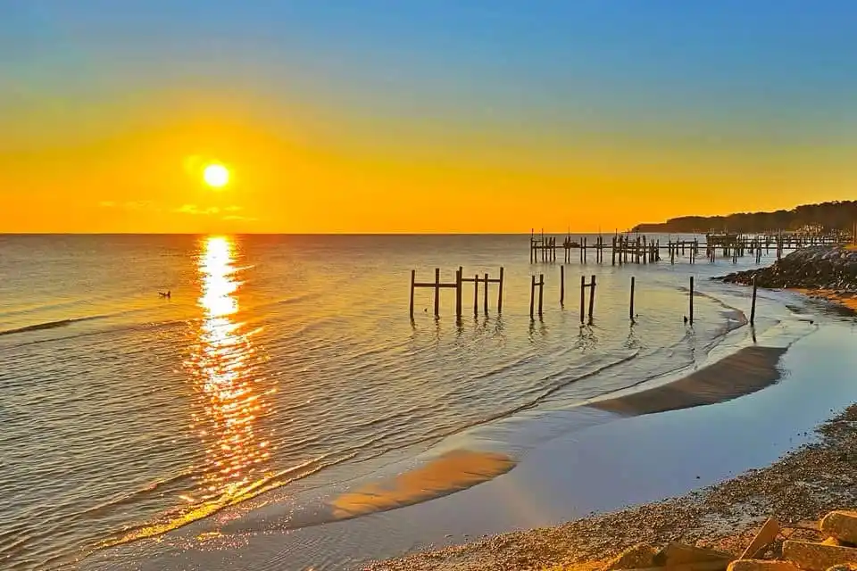 A serene sunset over a calm ocean with wooden pilings sticking out of the water near a pebbly shore. The sun is low on the…