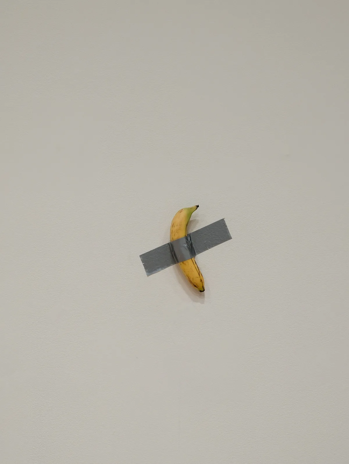 A single ripe yellow banana is affixed to a plain, light-colored wall with two strips of gray duct tape crossing over it in…